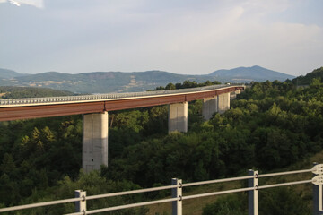 Obraz premium Side view of automobile bridge with highway road standing over green valley in countryside at evening in Europe. Soft focus. Copy space. Transportation building theme.