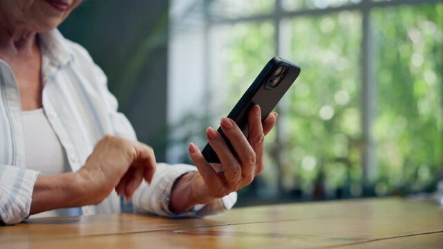 hands close-up of a very elderly gray-haired grandmother in a light blouse with a slight smile sitting against the background of a blue wall with large indoor plants writing a message on the phone