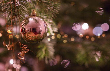 Golden Christmas ball hanging on fir tree against blurred festive lights.