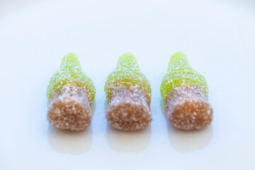 Detail studio shot of three small bottles of cola made of sugared gummy in a front view on a clean white plate. Concept of sweet colorful and luminous world, diabetes, whim and excess sugar