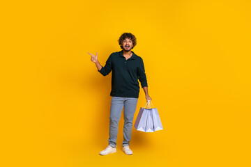 Young man with shopping bags pointing on yellow background, expressing excitement and fun. Ideal...