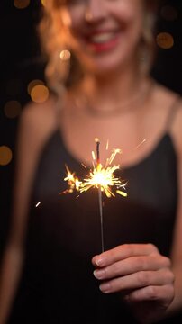 Christmas lights at the black background. woman blurry out of focus. New year eve celebration vibe. Slow motion vertical video footage. happy New year! dancing smiling blonde woman holding sparkler. 