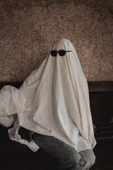 A whimsical figure draped in a white sheet, resembling a ghost, relaxes on a bench. Black sunglasses obscure its face, adding a humorous twist to the scene, evoking playful vibes.