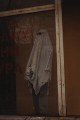 A ghostly figure draped in white stands behind a shop window, wearing dark sunglasses. The window features bold orange text, while the exterior shows a weathered facade
