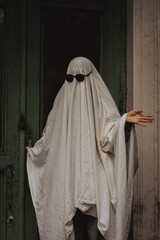 A playful figure draped in a white sheet mimics a ghost. Wearing dark sunglasses, it stands by an old green door, exuding a humorous, quirky vibe amidst urban surroundings.