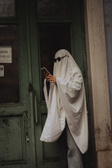 A person in a white sheet resembling a ghost stands outside a green door, reading a book. They wear sunglasses, creating a whimsical, mysterious vibe in a muted urban setting.