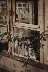 A spooky figure, draped in a white sheet, peeks through a rustic window adorned with vines....