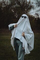 A person in a white ghost costume with sunglasses poses playfully in a forested area, surrounded by trees with autumn leaves and a moody sky, creating a whimsical yet eerie atmosphere. © Bohdan Hroshev
