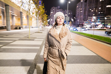 A woman in a light coat and a warm hat walks along a busy street in the evening city.