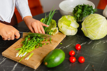 Close-up of hands slicing green onions on a wooden cutting board with a kitchen knife.
Fresh ingredients like lettuce and avocado in the background create a healthy cooking atmosphere.