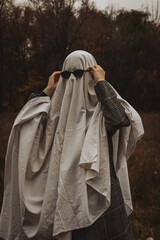 A figure draped in a flowing white fabric stands in a dimly lit field surrounded by trees, wearing...