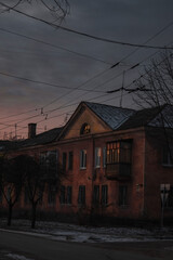 a twilight scene of a vintage, faded pink building with a balcony, warm light glowing from a window, surrounded by bare trees and tangled power lines against a dusky sky