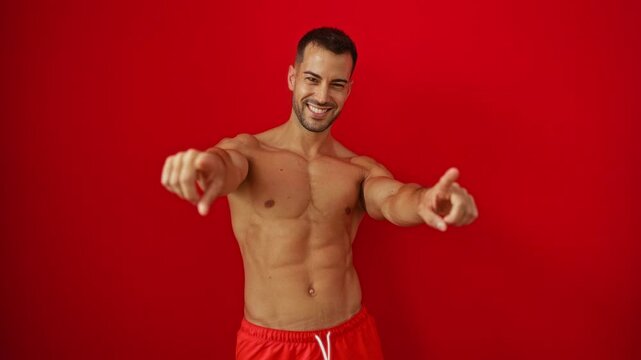 Young hispanic man shirtless in swimsuit pointing at you with a cheerful positive smile, finger pointing towards the camera over an isolated red background
