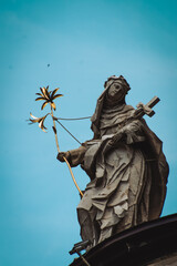 A tall statue of a woman holding a cross and a flower sprig stands atop a building against a vibrant blue sky, with fluffy white clouds providing a soft contrast.