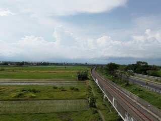 Railway tracks run between vast rice fields and a highway under a cloudy sky, capturing the harmony of nature and infrastructure.