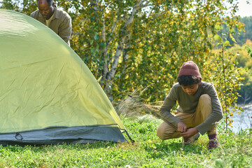 Boy squatting on grass while helping his father with pitching tent on hill, he trying to tie guy...