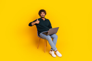 Young man with laptop giving a thumbs up gesture sitting casually on a chair against a vibrant yellow backdrop