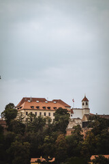 A majestic castle sits atop a hill, surrounded by lush green trees. Its red-tiled roof and white walls contrast against a cloudy sky, giving an air of historic elegance.