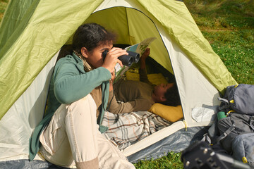 Mixed race teen girl sitting in tent and using binoculars while her little brother resting in tent and reading map