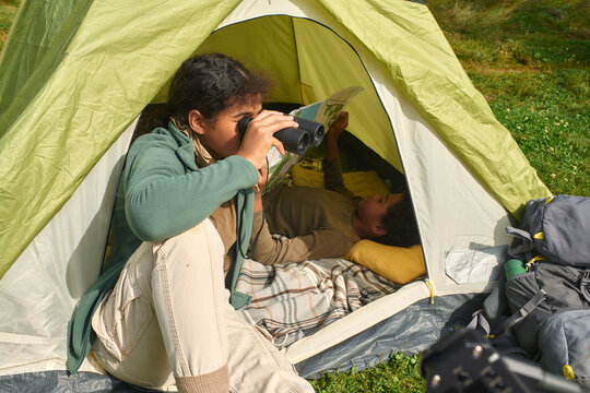 Mixed race teen girl sitting in tent and using binoculars while her little brother resting in tent and reading map