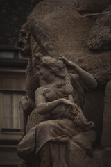 A detailed stone statue depicts a graceful figure with flowing robes, holding a textured object. The intricate craftsmanship captures emotion and elegance, set against a muted architectural backdrop.