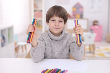 Portrait of cute boy with colorful pencils at white table in kindergarten