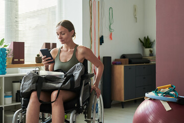 Woman sitting in wheelchair and looking at phone while in fitness center, gym equipment visible in...