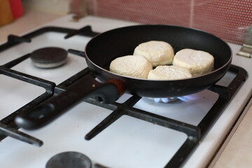 Raw syrniki (traditional cottage cheese pancakes) frying on a gas stove in a black non-stick pan. The soft white texture of the uncooked syrniki contrasts with the dark pan