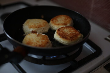 Golden-brown cheese pancakes frying in a black pan on a gas stove. A retro-styled teapot is visible in the background, creating a cozy and homely atmosphere