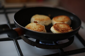 Golden-brown cheese pancakes frying in a black pan on a gas stove. A retro-styled teapot is visible in the background, creating a cozy and homely atmosphere