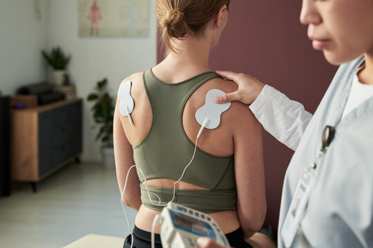 Woman experiencing therapeutic session with electrodes on back monitored by professional using specialized equipment in clinical setting focused on muscle stimulation