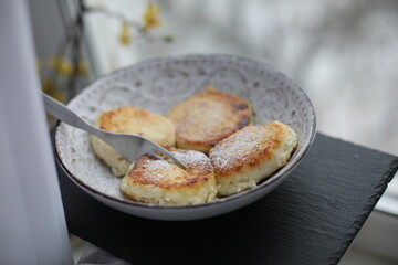 A plate of golden-brown syrniki (cottage cheese pancakes) dusted with powdered sugar, served with a fork on a patterned ceramic dish