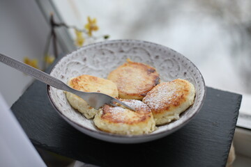 A plate of golden-brown syrniki (cottage cheese pancakes) dusted with powdered sugar, served with a fork on a patterned ceramic dish