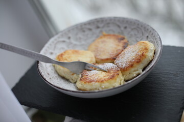 A plate of golden-brown syrniki (cottage cheese pancakes) dusted with powdered sugar, served with a fork on a patterned ceramic dish
