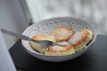 A plate of golden-brown syrniki (cottage cheese pancakes) dusted with powdered sugar, served with a fork on a patterned ceramic dish
