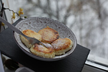 A plate of golden-brown syrniki (cottage cheese pancakes) dusted with powdered sugar, served with a fork on a patterned ceramic dish
