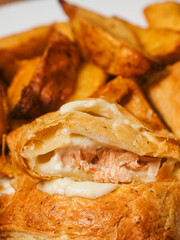 Close up of two crispy cooked salmon pie and golden wedges on a white plate and wooden table. Tasty simple dinner serving. Fried fine products. Pastry filled with a fillet of a fish and creamy sauce
