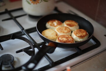Golden-brown cheese pancakes frying in a black pan on a gas stove. A retro-styled teapot is visible in the background, creating a cozy and homely atmosphere