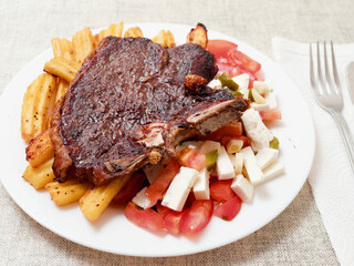 Dinner with premium t bone steak, fried potatoes chips with ridge, fresh salad with feta cheese on white plate and light color table cloth. Large portion of food with high quality products.