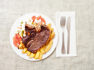 Dinner with premium t bone steak, fried potatoes chips with ridge, fresh salad with feta cheese on white plate and light color table cloth. Large portion of food with high quality products.