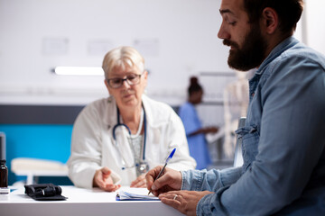 Closeup of young man filling out medical form in clinic and receiving patient guidance from caring senior doctor. Elderly female physician focused on male patient writing his information on clipboard.