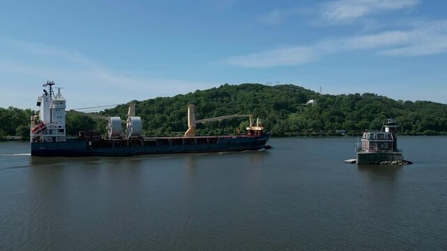 Large cargo ship sailing in a harbor with mountains in the background past a historic lighthouse, drone 4K