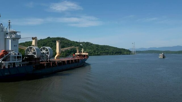 Large cargo ship sailing in a harbor with mountains in the background past a historic lighthouse, drone 4K