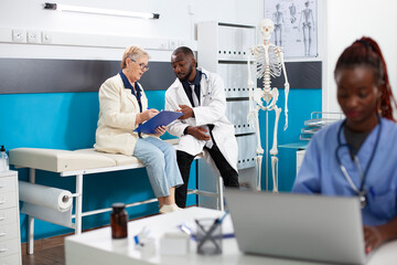 African american doctor and white old woman seated on hospital bed, reading medical information on...