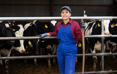 Young female farmer in overalls posing while feeding cows at dairy farm © JackF