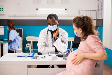 Expecting mother sits by office desk consulting with her doctor while female nurse uses desktop pc in background. Pregnant white woman and male obstetrician with face masks reviewing health documents.