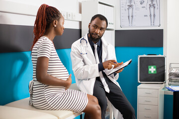 African american doctor sits next to pregnant lady while showing medical results on device. Black woman with hand on pregnancy belly listens to maternal advice from physician holding digital tablet