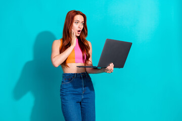 Surprised young woman with red hair wearing summer casual outfit holds laptop against a vibrant teal background