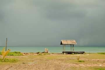 A wooden gazebo by a calm beach, surrounded by the sea beneath cloudy skies, creates a peaceful...