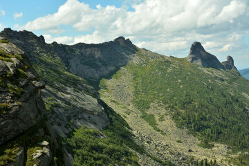 A steep slope of a high mountain with a narrow ridge in a picturesque valley under a summer cloudy sky.